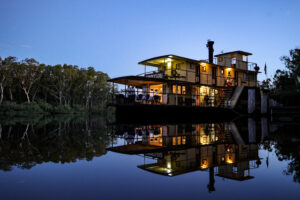 Evening photo of PS Emmylou paddle steamer illuminated and reflected in the calm waters of the Murray River, surrounded by gum trees under a soft twilight sky.