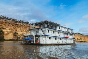 The Proud Mary riverboat cruising along the Murray River in front of sunlit ochre cliffs and a wide blue sky.